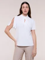 Blusa blanca de corte recto con cuello alto torzado, abertura frontal en gota y mangas cortas con aberturas sobre los hombros.