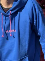 Canguro de tela deportiva color azul con capucha y cordones ajustables en color rosa. Presenta el logo bordado en color rosa en el pecho.