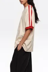 Remera oversize de color crudo con escote en V y cuello en rib rojo, mangas cortas con rib rojo y una franja roja en los hombros. Cuenta con un estampado frontal de bailarinas y detalles en relieve.
