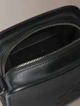 Cartera tipo reporter de Calvin Klein confeccionada en piel sintética suave de color negro. Presenta un diseño cuadrado con monograma tonal grabado en el frente, correa ajustable para cruzar, cierre de cremallera superior y bolsillo exterior trasero.