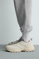 Zapatilla deportiva tipo running con diseño de paneles superpuestos en combinación de materiales. Presenta cordonera curvada y suela con volumen de diseño irregular.