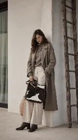 Cartera tipo bandolera confeccionada en cuero vacuno con pelo (nonato), con diseño bicolor en tonos marrón oscuro y blanco. Presenta costuras artesanales en los laterales, cierre central con detalle de botón y correa larga ajustable.