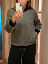 Chaqueta tipo bomber de lana con estampado de espiga en tonos grises. Presenta cuello, puños y ruedo en tejido elástico negro, cierre frontal dorado y bordado sutil en el pecho.