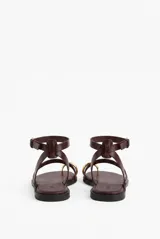 Sandalias planas de cuero color negro, con tiras finas, sujetador para el dedo y detalle de anilla metálica en la puntera. Cuenta con tira ajustable alrededor del tobillo con hebilla plateada.