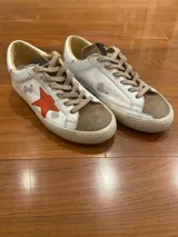 Zapatillas bajas de cuero vacuno color blanco con detalles gastados y patinados en gris. Aplique de estrella roja en el lateral externo, puntera de gamuza visón y talón superior de cuero plateado. Cordones de algodón encerado y suela de caucho.