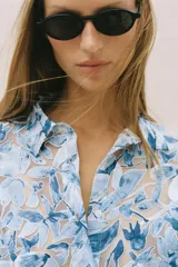 Camisa de manga larga con cuello solapa y cierre frontal de botones, confeccionada en tejido combinado semitransparente con estampado floral en tonos azules y blancos.