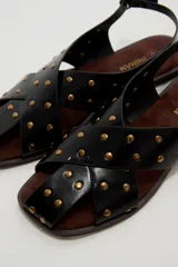 Sandalias planas de cuero sintético marrón, con tiras cruzadas en el empeine y tachas metálicas doradas distribuidas por toda la capellada. Cuenta con tira ajustable en el talón con hebilla.