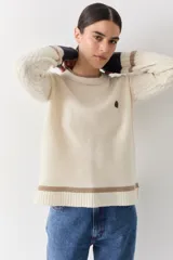 Buzo de punto color crudo con mangas de textura trenzada, detalles en contraste en los puños y franjas horizontales en el ruedo. Presenta cuello redondo y un pequeño bordado en el pecho.