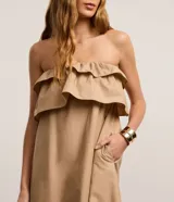 Vestido largo color beige, estilo evasé, confeccionado en tricolina de algodón. Presenta escote hombro a hombro con un volante superpuesto y bajo fruncido.