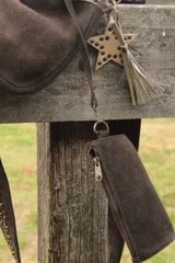 Bandolera confeccionada en cuero de gamuza marrón, con cierre superior. Presenta un llavero colgante con forma de estrella revestido en tachas metálicas y tiras flecadas en tono dorado.