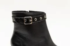 Botas de cuero negro, de caña baja, punta fina y taco cuadrado. Presentan un aplique charolado al tono, una correa con hebilla y ojales metálicos.
