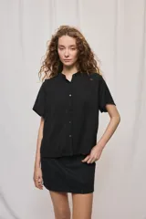 Camisa negra de manga corta, silueta amplia y tejido liviano con textura. Presenta cuello mao, cierre frontal con botones y diseño fluido.