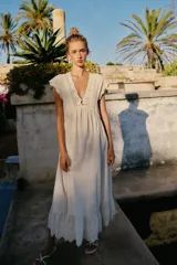 Vestido largo color crudo, sin mangas, con escote en V pronunciado, canesú superior de tejido de punto con botones frontales y lazo para atar en el cuello, y falda fluida con detalle de bordado en el bajo.