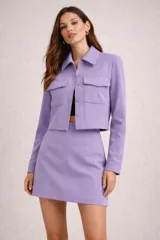Conjunto de chaqueta y minifalda color lila, de corte sastre. La chaqueta cuenta con cuello camisero, cierre frontal de cremallera y botones, bolsillos con solapa en el pecho y diseño corto. La falda es de corte recto y tiro alto.