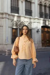 Blusa de manga larga con estampado de cuadros vichy en tonos amarillo y rosado. Presenta cuello mao con escote en V y botones frontales. Las mangas son amplias con puños terminados en volados fruncidos.
