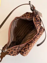 Cartera tipo baguette con estampado animal print, confeccionada en material suave símil gamuza. Presenta un bolsillo frontal con solapa y cierre, compartimento principal con cierre superior, y asa larga regulable de cuero sintético marrón.