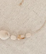 Collar de diseño artesanal compuesto por piedras irregulares en tonos crudos veteados, con una pieza central de mayor tamaño. Presenta un cordón beige ajustable y detalles en tono dorado.