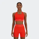 Top deportivo de soporte medio, color naranja brillante, con escote redondo y espalda tipo nadador. Presenta una banda elástica ancha bajo el busto con el logo de New Balance repetido en blanco.