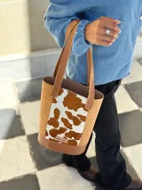 Bolso tipo matera confeccionado en cuero, con diseño combinado que presenta laterales con textura de cocodrilo y panel central con estampado animal print de vaca. Cuenta con asas largas para llevar al hombro y base reforzada.