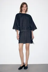 Vestido corto de denim azul oscuro, con cuello redondo, mangas amplias de largo tres cuartos y cintura elástica con cinturón integrado del mismo tejido. Presenta costuras a contraste y cierre posterior con cremallera oculta.