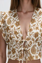 Blusa de diseño estampado con motivos florales y cachemira en tonos marrones sobre base blanca, con escote en V profundo y doble lazo frontal ajustable. Presenta mangas cortas con volados.