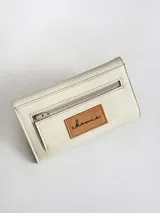 Billetera de cuero vacuno color crudo con textura granulada. Presenta un diseño tipo sobre con cierre de broche y un detalle decorativo de espiga bordado en cuero color suela en la solapa frontal.
