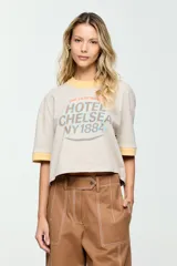 Remera de corte amplio, color beige, con mangas cortas y cuello redondo. Presenta un estampado frontal con la inscripción "The Legendary Hotel Chelsea NY 1884" en tonos celeste y marrón, con detalles en amarillo en el cuello y los puños.