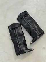 Botas texanas de caña alta en color negro, confeccionadas en cuero sintético. Presentan un diseño con cordones frontales a lo largo de toda la caña, rematados con un lazo en la parte superior, y puntera metálica plateada.