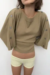 Jersey de punto color beige con cuello barco y mangas anchas tipo capa que presentan detalle de botones a lo largo de los hombros y brazos.
