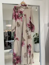 Vestido largo beige de corte camisero con estampado floral en tonos fucsia y marrón, cuello camisero, cierre frontal con botones y mangas largas.