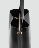 Bolso tipo saca de diseño minimalista en color negro, con asa de hombro fija y detalles de botones metálicos dorados en los laterales. Cuenta con cierre de mosquetón y forro interior.