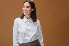 Camisa blanca de corte clásico, con cuello camisero, mangas largas con puños abotonados y cierre frontal con botones decorativos en forma de estrella con brillo.