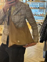Cartera tipo hobo de gamuza color avena con diseño amplio y costuras decorativas. Cuenta con correa ajustable de cuero marrón y una placa metálica dorada con el logo de la marca en la parte inferior.