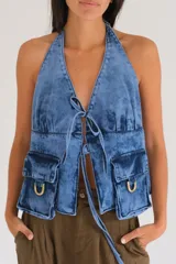 Top halter de denim azul con efecto deslavado, escote pronunciado, cierre frontal con tiras para anudar, bolsillos cargo delanteros con detalles de estribos metálicos y diseño utilitario.