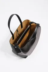 Bolso de hombro tipo tote bag de piel vacuna color beige, con doble asa de mano y correa ajustable de tejido. Presenta un cierre de cordón en la parte superior y un logo metálico en relieve en el frente. Incluye bolsillo interior con cierre de cremallera.