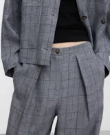 Pantalón de lino gris con estampa de cuadros, tiro medio y pierna ancha. Presenta pliegues frontales, cierre con botón y elástico en la parte posterior de la cintura.