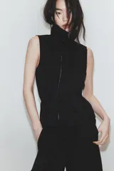 Chaleco negro de diseño fluido con cuello subido y cierre frontal de cremallera metálica. Presenta cordones ajustables con lazadas en el cuello y en la cintura.