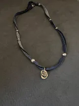 Collar de estilo artesanal compuesto por una cinta de tela en tonos azulados y grisáceos, adornada con pequeñas esferas metálicas plateadas. Presenta un dije central de metal con forma ovalada irregular que muestra el símbolo del Espíritu Santo en relieve.