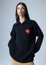 Sweater negro de cuello polo tejido con cierre diente de perro a tono y tajos laterales. Tiene un parche bordado con elementos del drop.