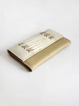 Billetera de cuero con diseño bicolor en tonos crema y beige. La solapa presenta una textura tipo reptil y detalles decorativos de cordones cruzados en los laterales. Cuenta con cierre mediante broche y monedero externo.