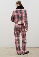 Pantalón de tiro alto confeccionado en denim de corte recto, con estampado artístico abstracto en tonos rosados, rojos y negros. Presenta cierre frontal con botones visibles.