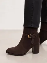 Bota de caña media confeccionada en cuero gamuzado color marrón oscuro, con punta almendrada y taco cuadrado de altura media. Presenta una tira decorativa con hebilla metálica dorada en el tobillo.