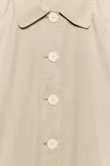 Trench largo de Zara Woman Collection, confeccionado en color beige. Cuenta con cuello de solapa, manga larga, bolsillos frontales de parche y cierre central con botones visibles.