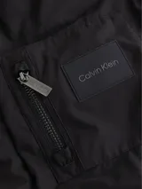 Campera bomber Calvin Klein negra, de corte regular, con cuello, puños y bajo de punto de canalé. Cierre frontal con cremallera, bolsillo con cremallera en la manga izquierda y bolsillos de hendidura en la cintura. Forro de malla e insignia con el logo de goma de Calvin Klein en la manga.