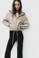 Campera estilo cazadora con capucha ajustable con cordones, cierre frontal de cremallera, mangas largas con puños elásticos, bolsillos delanteros de doble vivo y bajo ajustable con cordones. Presenta un diseño de cuadros en tonos beige y marrón.