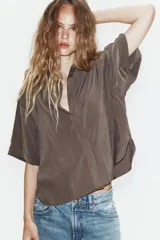 Camisa de corte fluido con cuello solapa y manga corta con dobladillo. Presenta un cierre frontal con botones ocultos bajo una tapeta y un diseño con bajo ligeramente más largo en la parte posterior.
