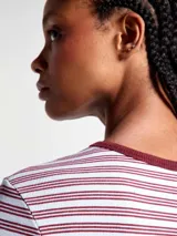 Camiseta de manga larga con cuello redondo, confeccionada en tejido acanalado con diseño de rayas horizontales en tonos claros y bordó. Presenta un pequeño bordado decorativo en el centro del pecho.