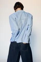 Camisa de manga larga con cuello solapa y hombreras. Presenta un diseño de rayas verticales en tonos azul y blanco, con cierre frontal de botones y puños anchos.