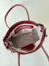 Cartera de cuero de tamaño medio con estructura rígida. Presenta una tira decorativa con tachas y hebillas metálicas en los extremos, además de lazos laterales. Cuenta con doble asa para llevar al hombro y cierre superior.