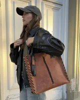 Cartera tipo tote de cuero marrón con diseño estructurado. Presenta detalles de trenzado lateral y central en color negro, con remaches metálicos decorativos. Incluye asas de hombro fijas y el logo de la marca grabado en la parte superior.